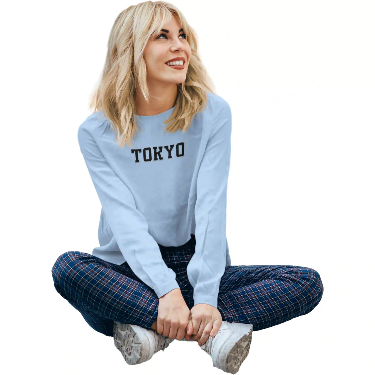 Woman wearing a light blue sweatshirt with 'TOKYO' text, sitting cross-legged on a white background.